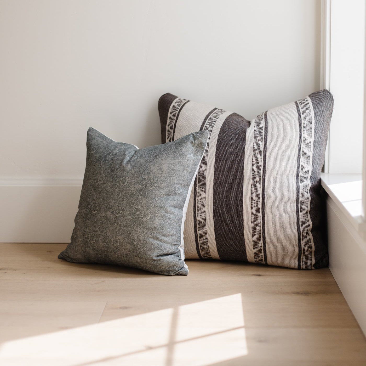 Two throw pillows in gray and cream with striped and geometric patterns on wooden floor against white wall.