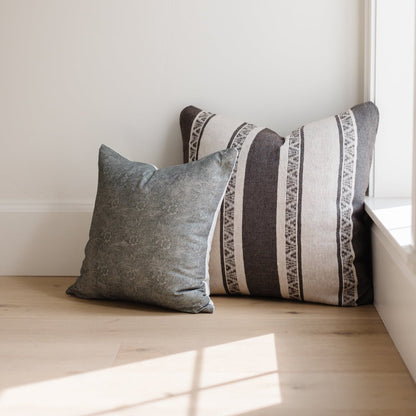 Two throw pillows in gray and cream with striped and geometric patterns on wooden floor against white wall.