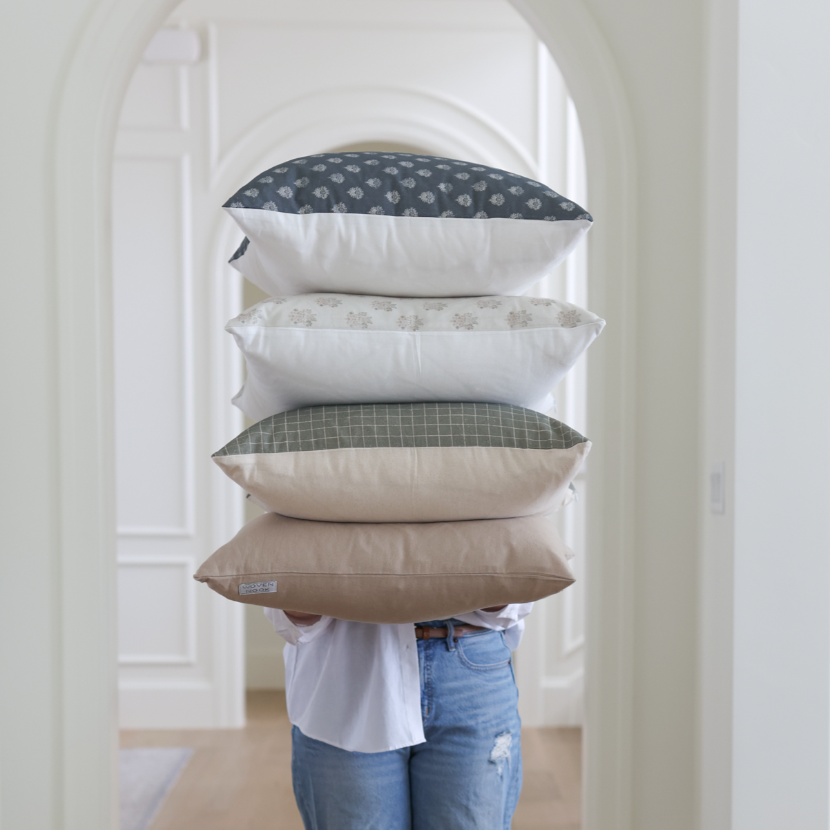 Five stacked throw pillows in neutral tones with various patterns, held by a person wearing a white shirt and jeans.