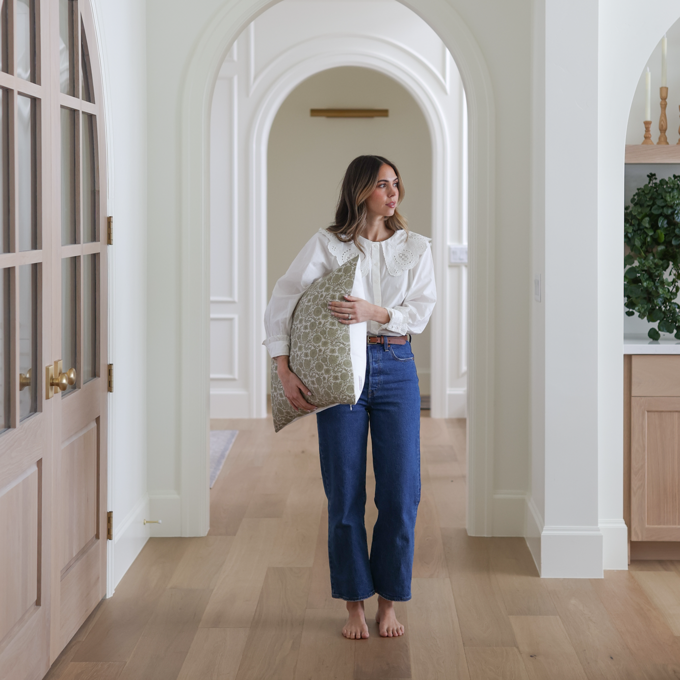 Woman holding a green and cream patterned pillow while standing in a hallway, wearing a white blouse and dark jeans.