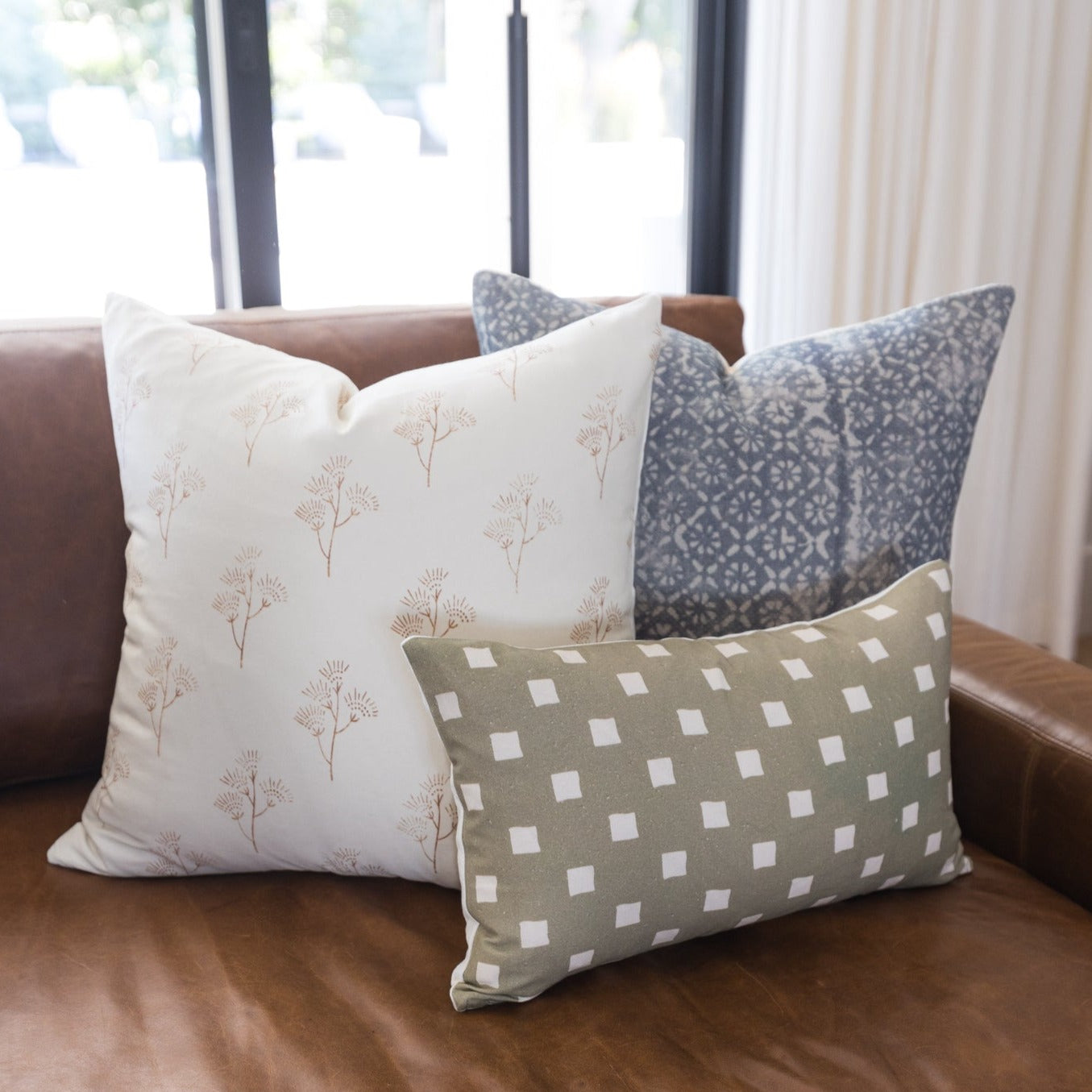 Three decorative pillows with floral and geometric patterns in cream, gray, and taupe on a brown leather sofa.