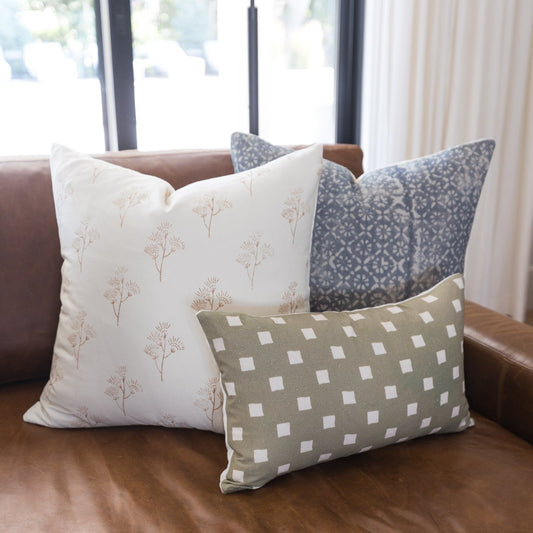Three decorative pillows with floral and geometric patterns in cream, gray, and taupe on a brown leather sofa.
