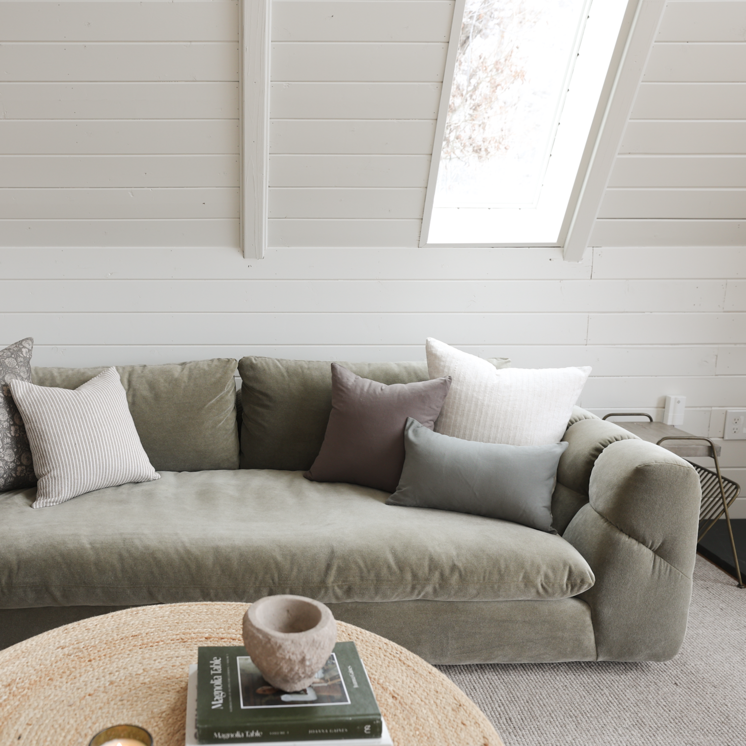 Beige upholstered sectional sofa with multiple throw pillows in neutral tones, displayed in a bright room with skylights.