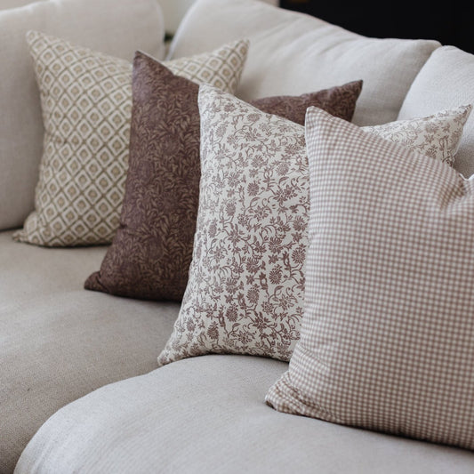 Four throw pillows in neutral tones with geometric, floral, and gingham patterns arranged on a light-colored sofa.