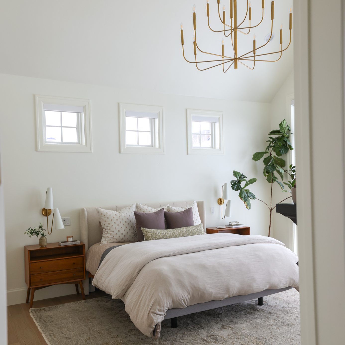 Gold-finished multi-arm chandelier with cylindrical shades hanging above a bedroom with bed, nightstand, and windows.