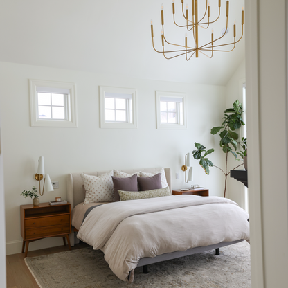 Gold-finished multi-arm chandelier with cylindrical shades hanging above a bedroom with bed, nightstand, and windows.