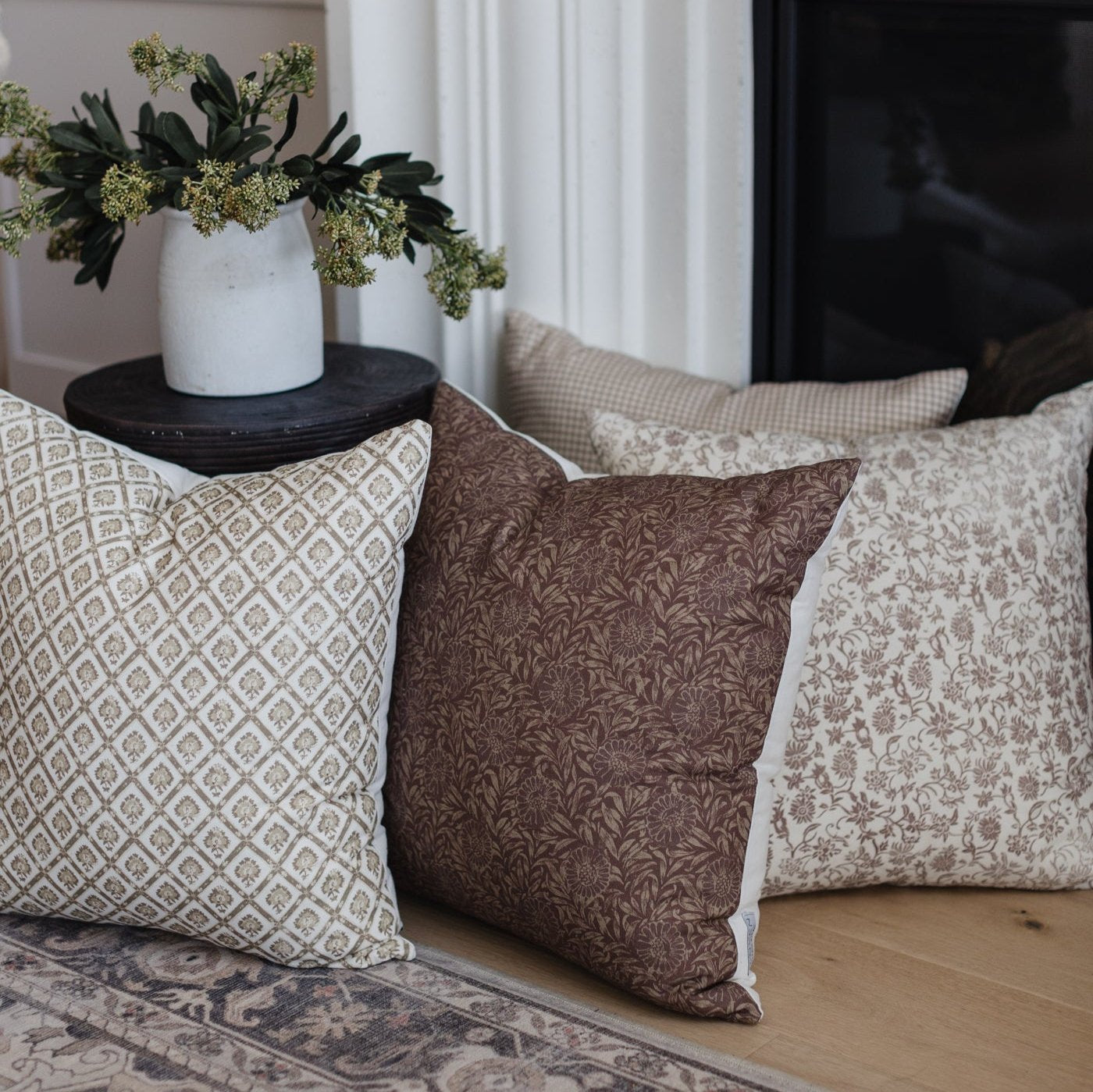 Three decorative pillows in neutral tones with geometric and floral patterns arranged on a wooden floor next to a side table