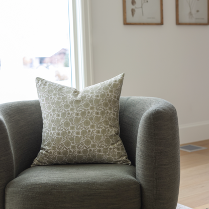 Green and cream patterned square pillow displayed on a gray upholstered armchair.