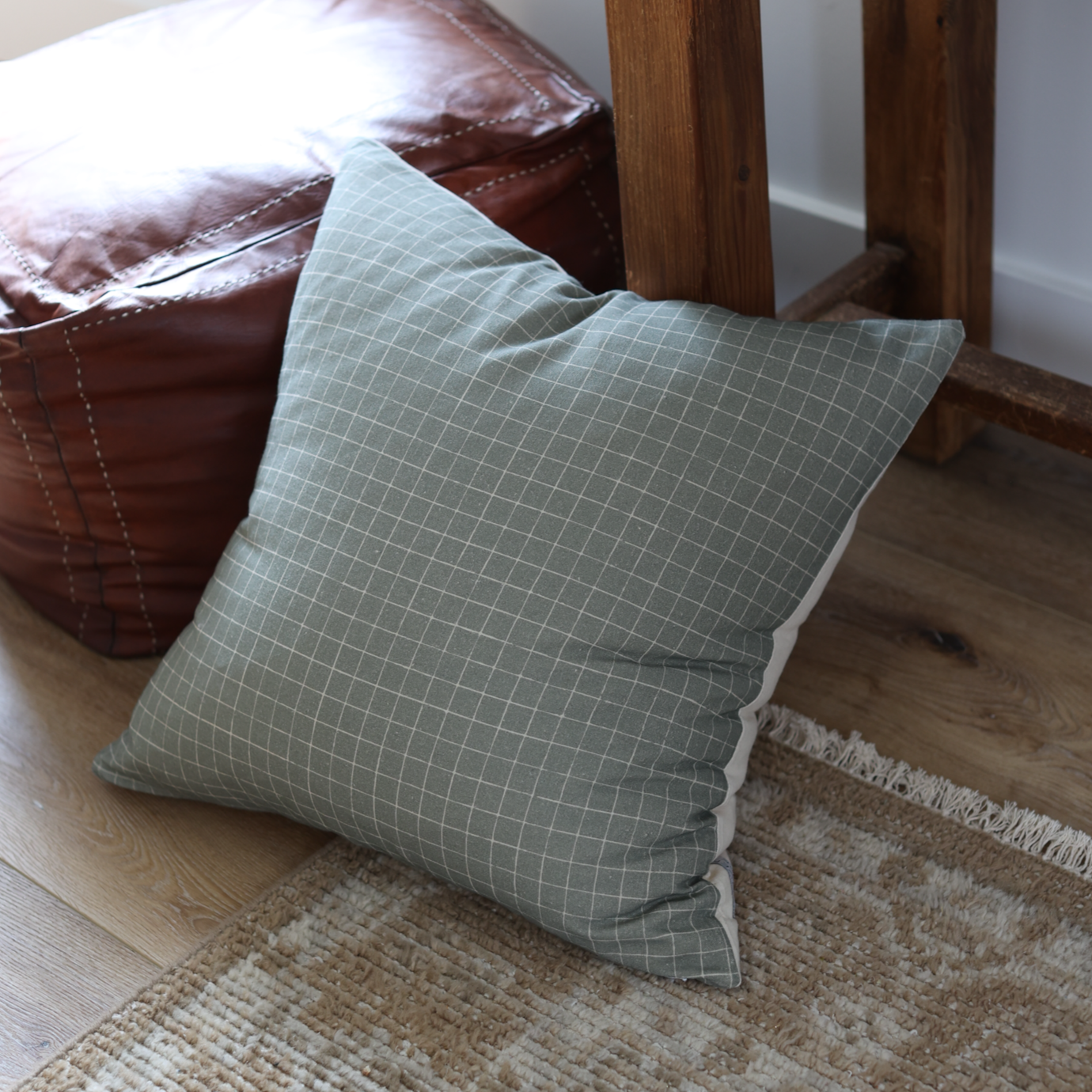 Moss green grid patterned pillow with white piping on wooden chair next to brown leather furniture.