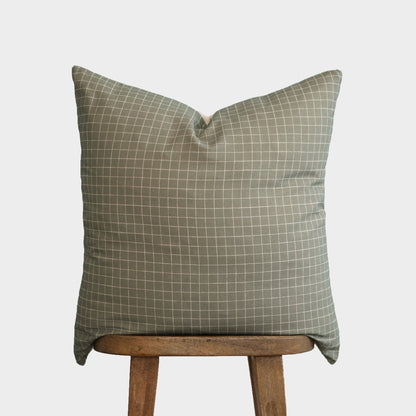 Moss-colored square pillow with white grid pattern displayed on wooden chair leg.