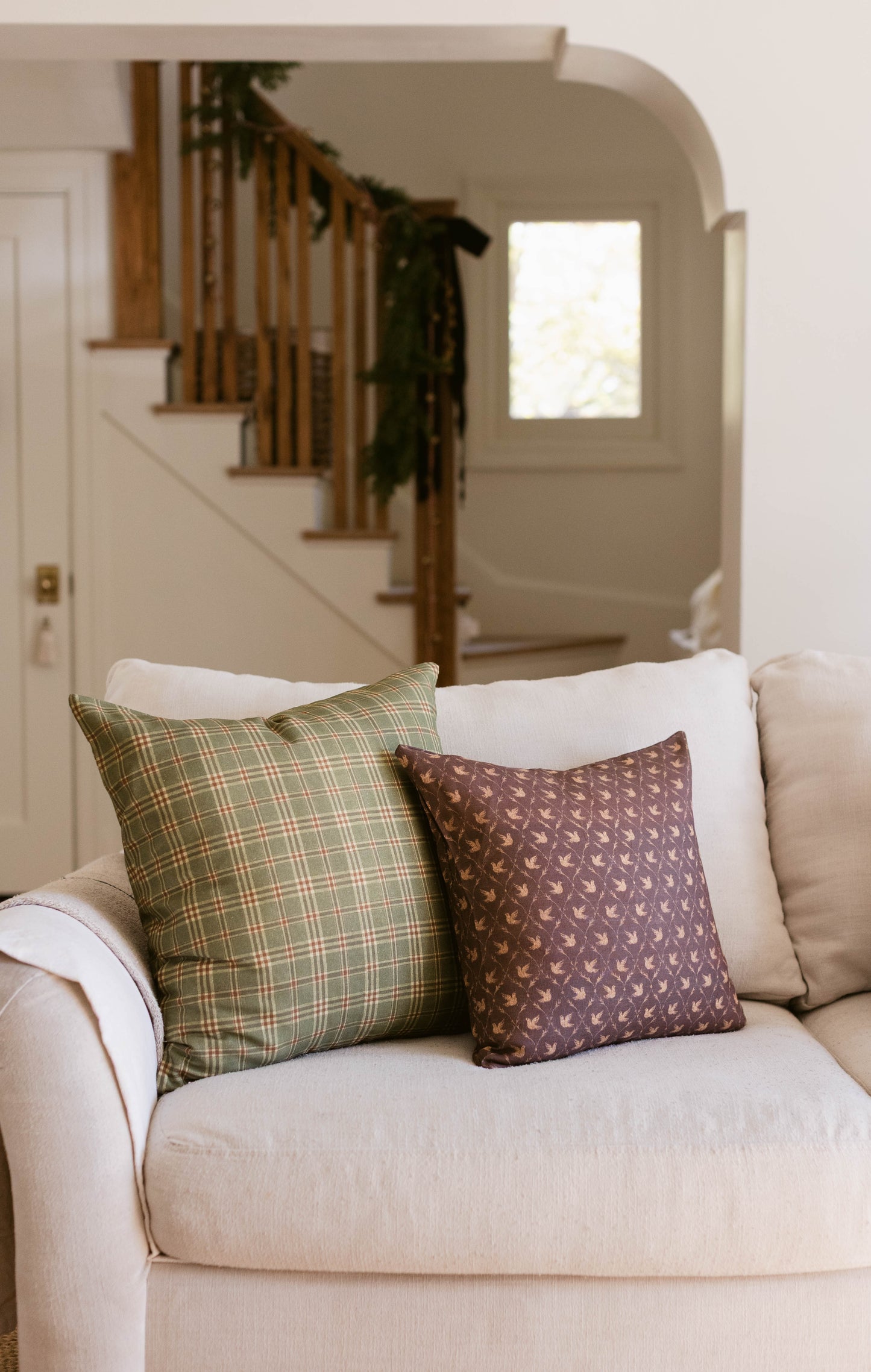 Cream upholstered sofa with green plaid and brown patterned throw pillows in a residential entryway.
