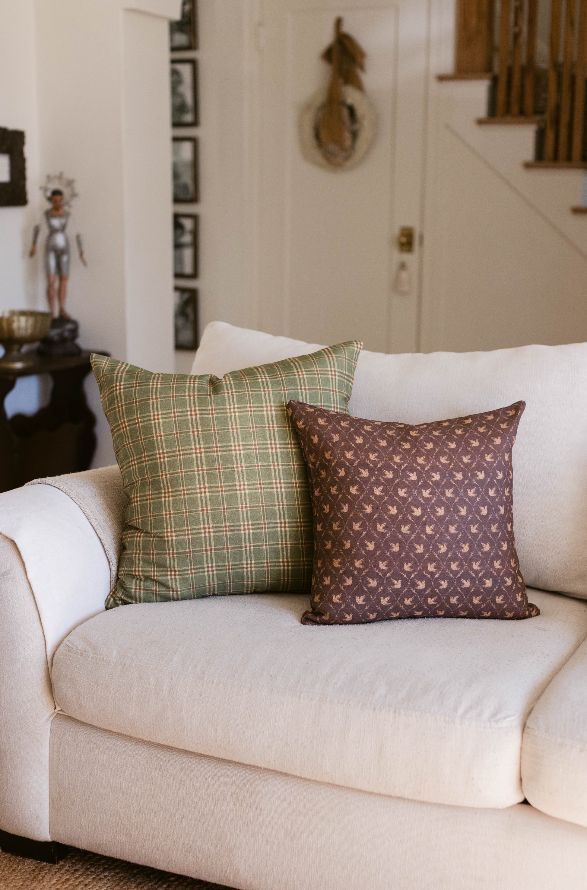 Two decorative pillows on a cream upholstered chair: one with green plaid pattern, one with brown bird motif.