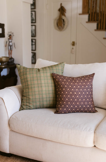 Two decorative pillows on a cream upholstered chair: one with green plaid pattern, one with brown bird motif.