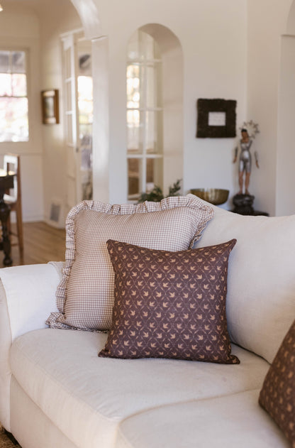 Brown patterned pillow with bird motifs on cream sofa next to checkered and white pillows.