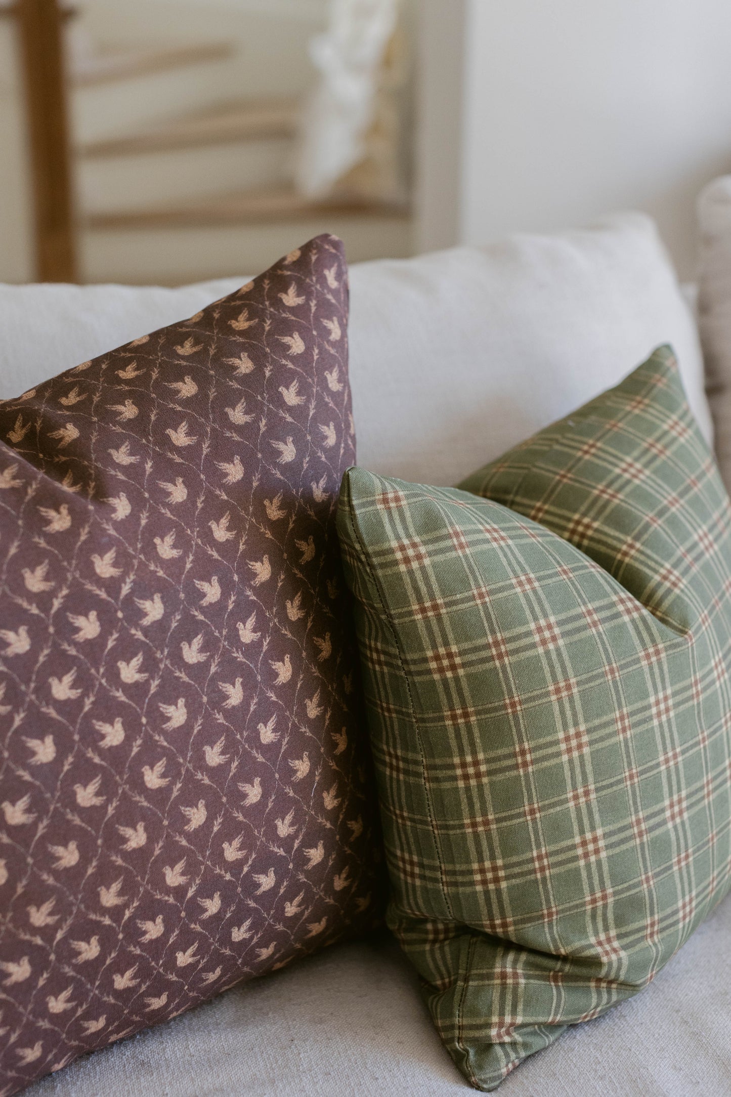 Purple bird-patterned pillow and green plaid pillow arranged on white bedding with wooden furniture in background.