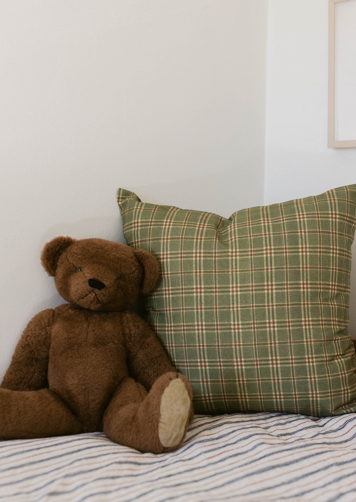 Green plaid pillow with cream and red stripes displayed next to brown teddy bear on striped bedding.