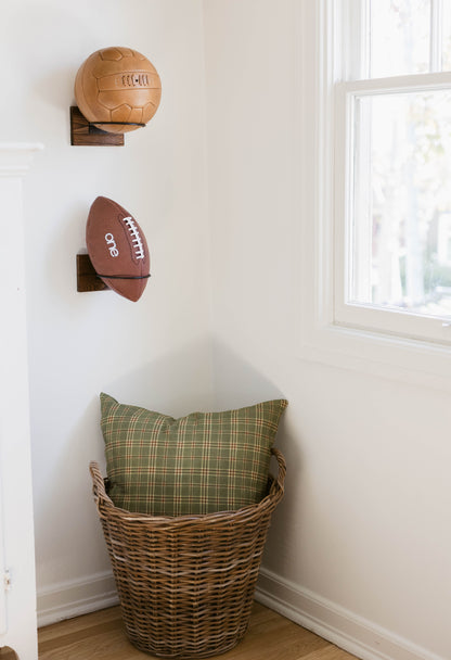Tan leather soccer ball and brown football mounted on wooden wall brackets above wicker basket with green plaid pillow.