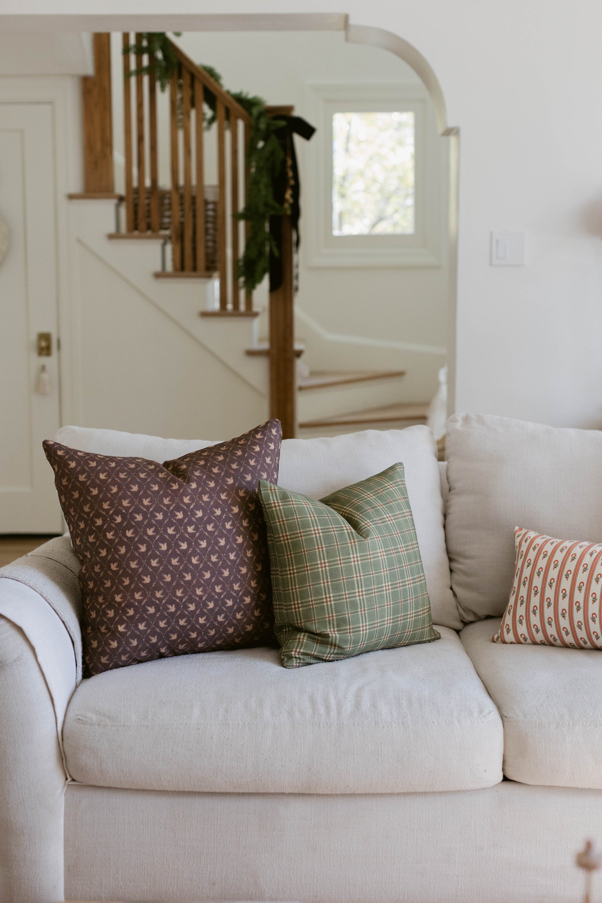 Decorative pillows in brown, green plaid, and coral patterns arranged on a cream upholstered sofa.