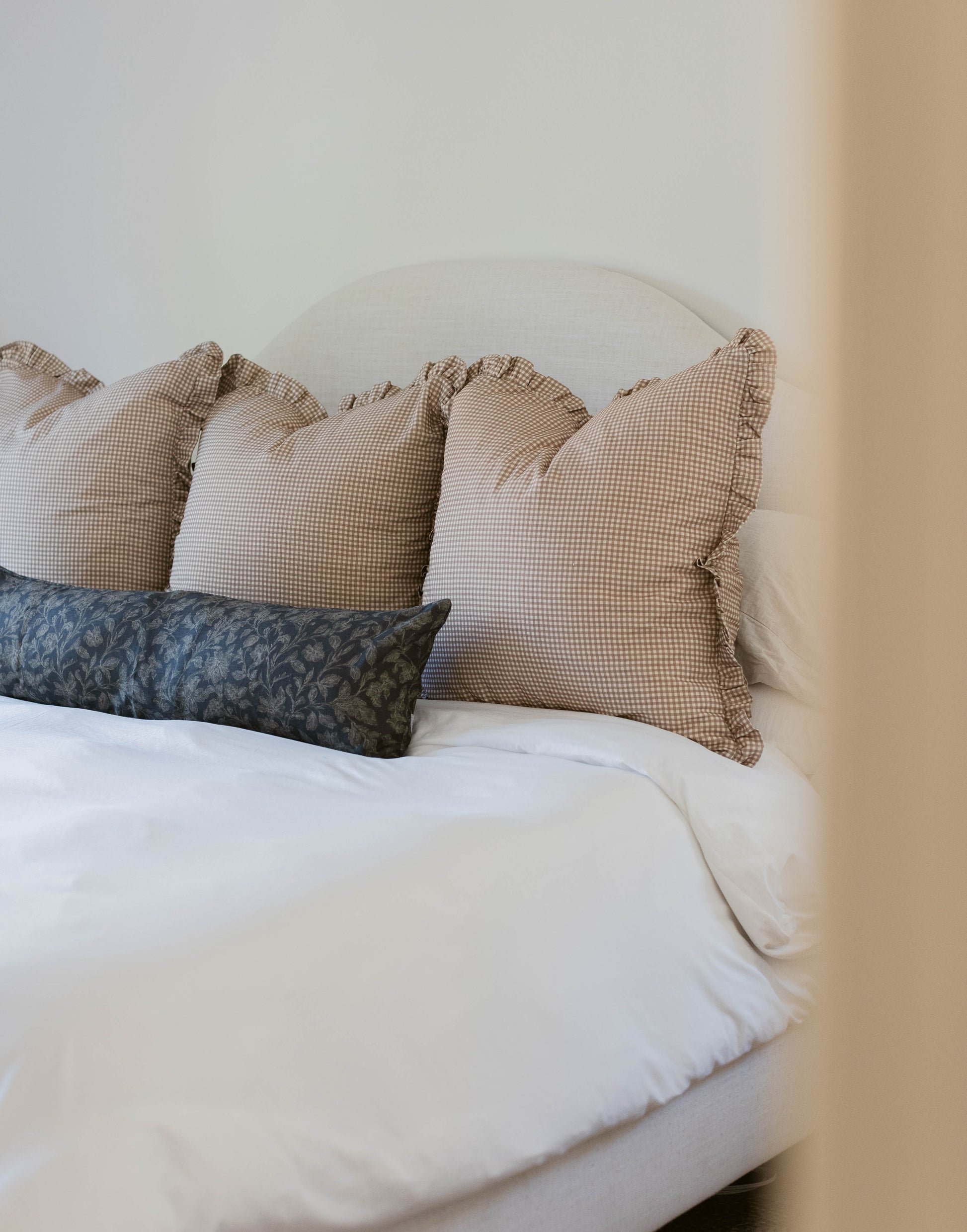 Tan and white gingham pillows with ruffled edges arranged on a bed with white linens.