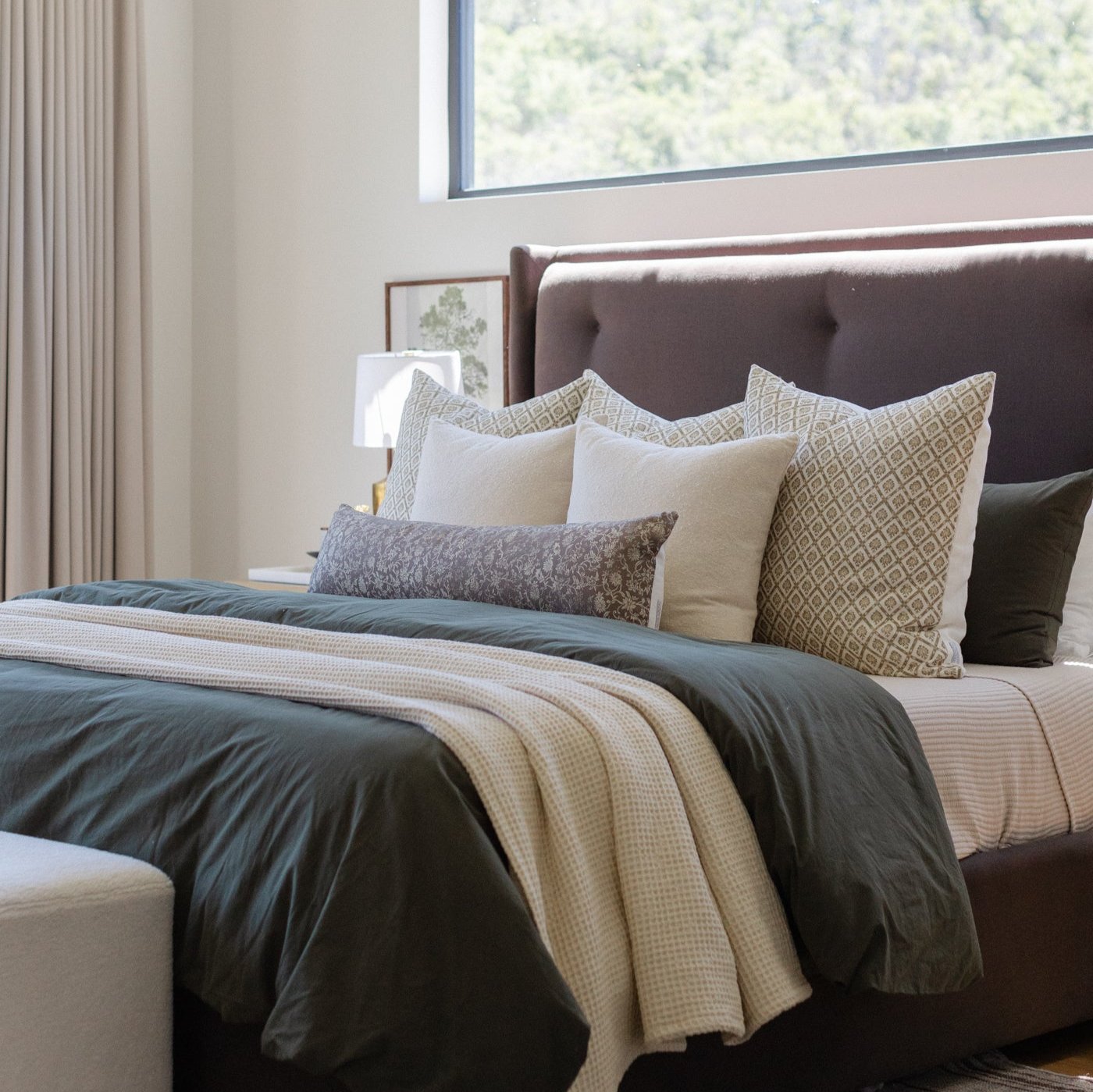 Bed styled with dark charcoal duvet, cream and tan patterned linen pillows, and gray lumbar pillow against brown upholstered 