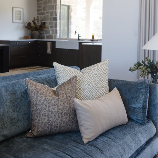 Three decorative pillows on a blue sofa: one with beige damask pattern, one with geometric diamond print, and one solid cream