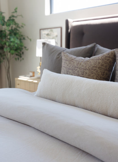 Off-white boucle lumbar pillow with textured weave displayed on a bed with gray pillows.
