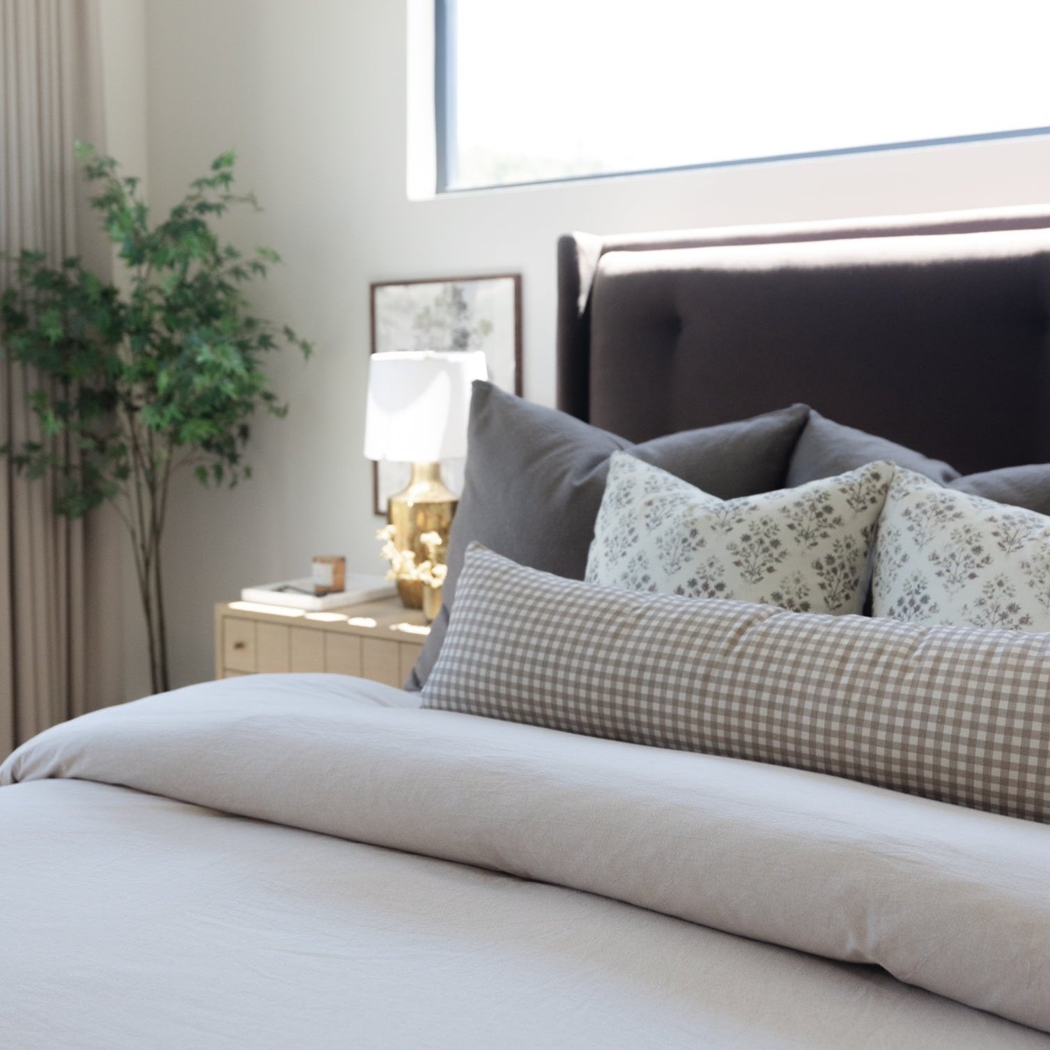 Long beige pillow with solid fabric displayed on bed with gray checkered and patterned pillows.