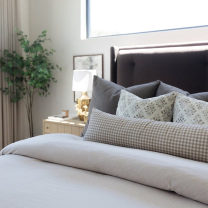 Long beige pillow with solid fabric displayed on bed with gray checkered and patterned pillows.