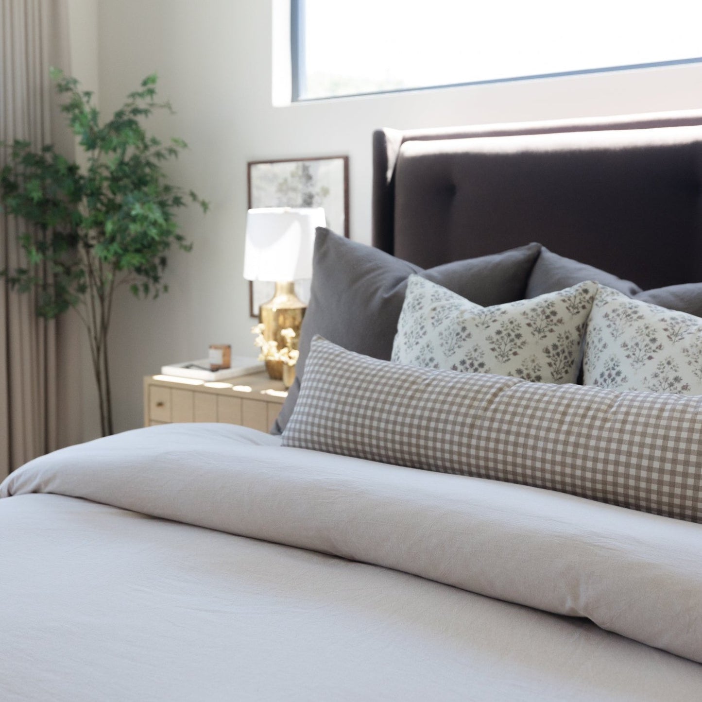 Tan and white gingham lumbar pillow displayed on a bed with gray pillows and white bedding.