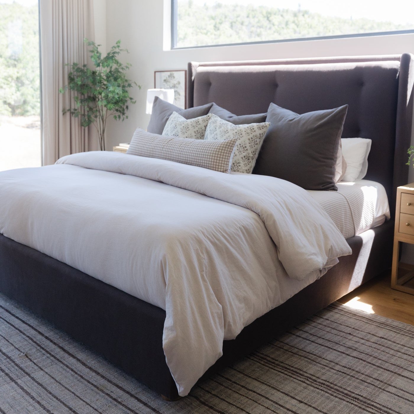 Dark gray upholstered bed frame with white bedding, decorative pillows, and wood nightstand in bright bedroom.