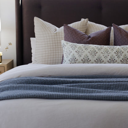 Lumbar pillow with cream gingham pattern displayed among coordinating decorative pillows on a bed with blue waffle-weave blan