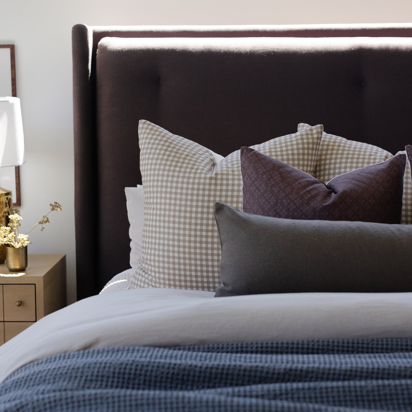 Tan and white gingham check decorative pillow displayed on a bed with dark headboard and layered bedding.