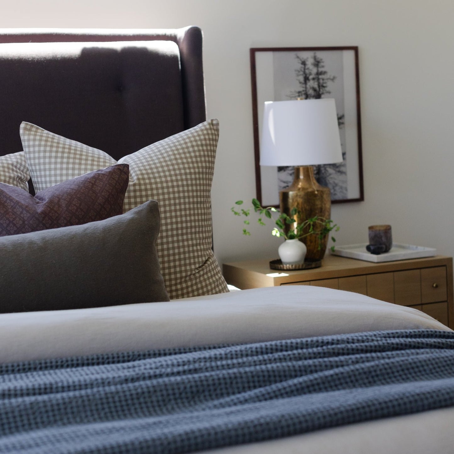 Tan and white gingham patterned pillow displayed on a bed with dark headboard and layered bedding.