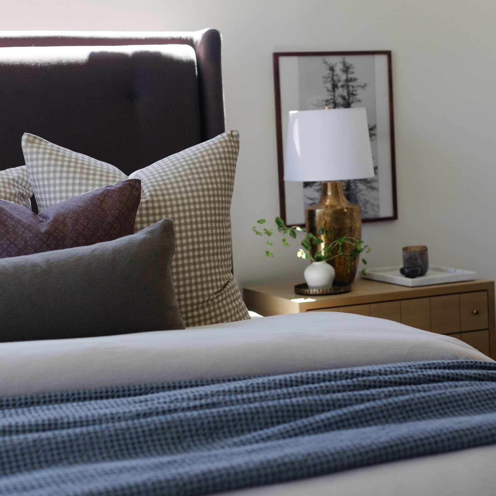 Tan and white gingham patterned pillow displayed on a bed with dark headboard and layered bedding.