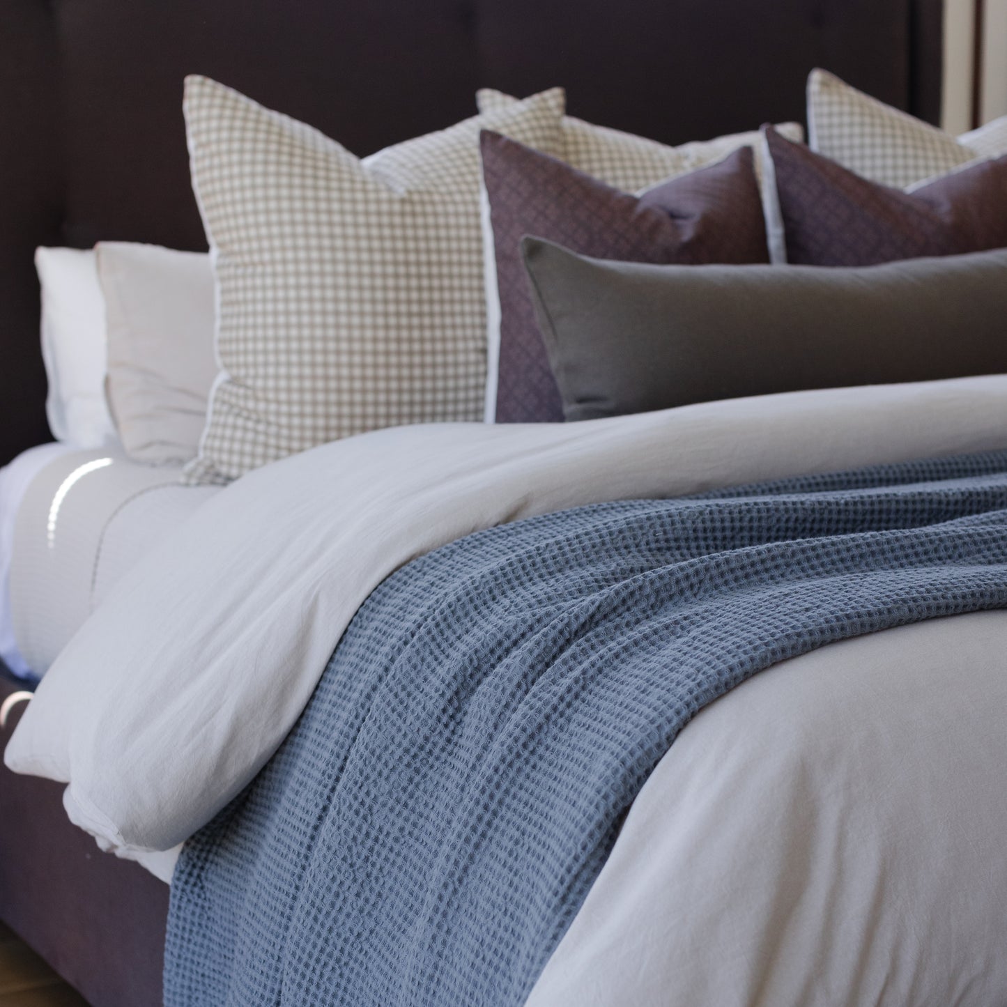 Tan and white gingham patterned pillow displayed on a bed with white sheets and blue throw blanket.