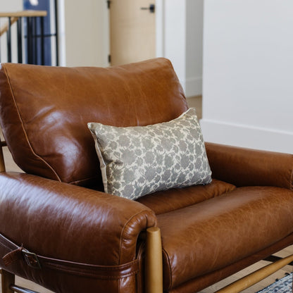 Gray floral patterned lumbar pillow displayed on a brown leather armchair with wooden frame.