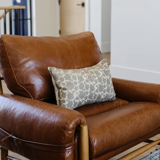 Gray floral patterned lumbar pillow displayed on a brown leather armchair with wooden frame.