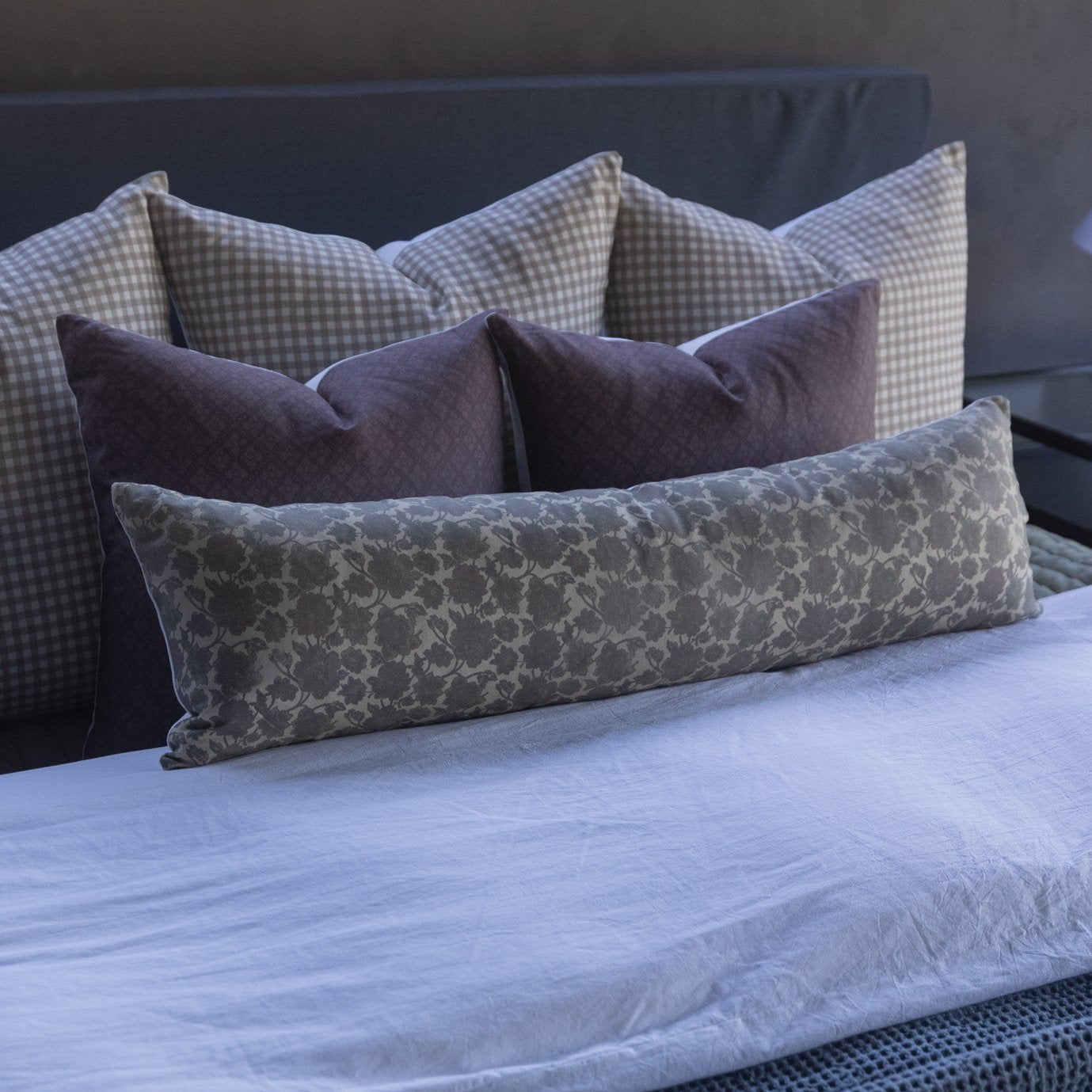 Maroon lumbar pillow arranged with gingham and patterned pillows on a bed with blue bedding.