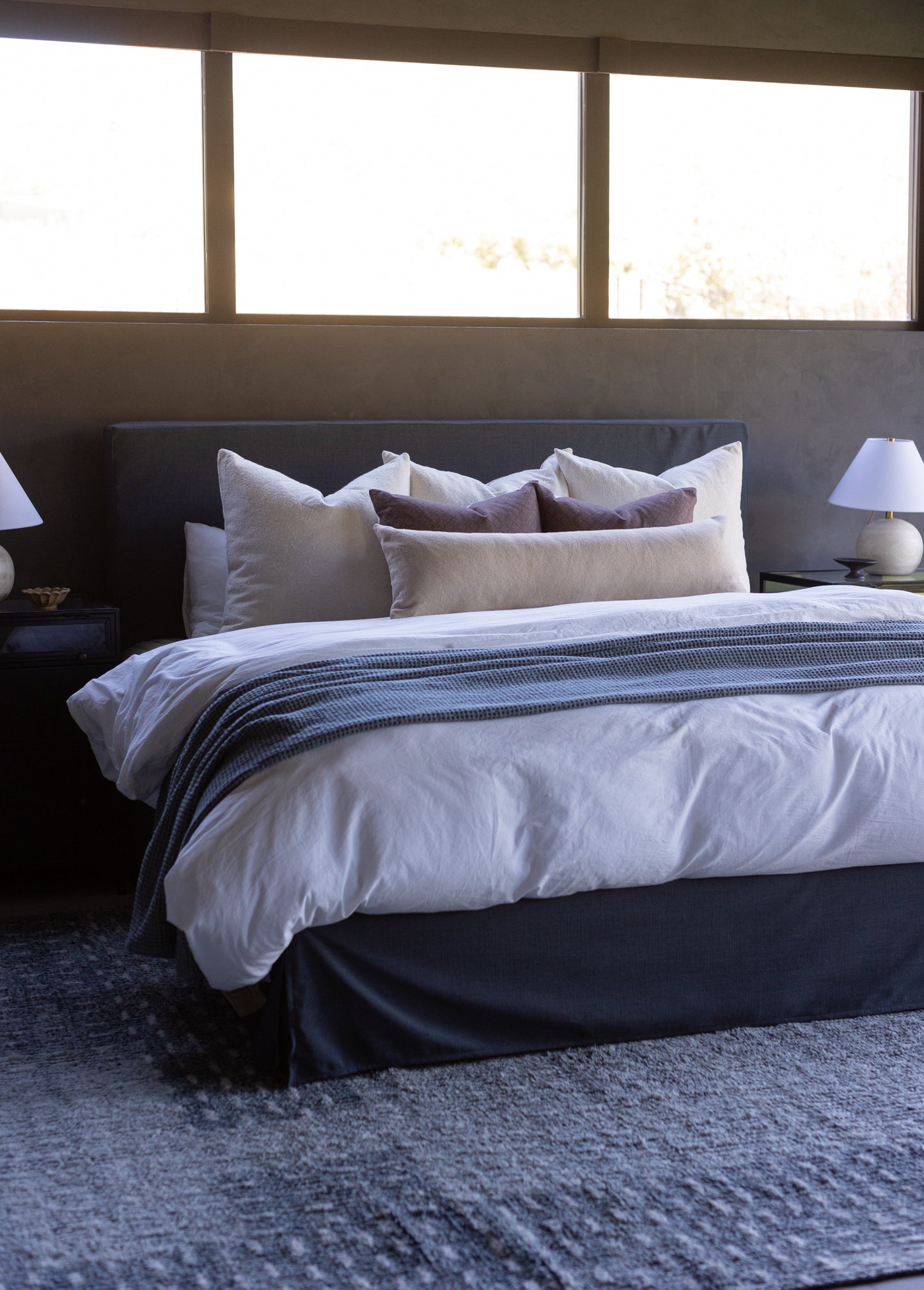 Beige boucle textured pillows arranged on a dark headboard with white bedding and striped throw.