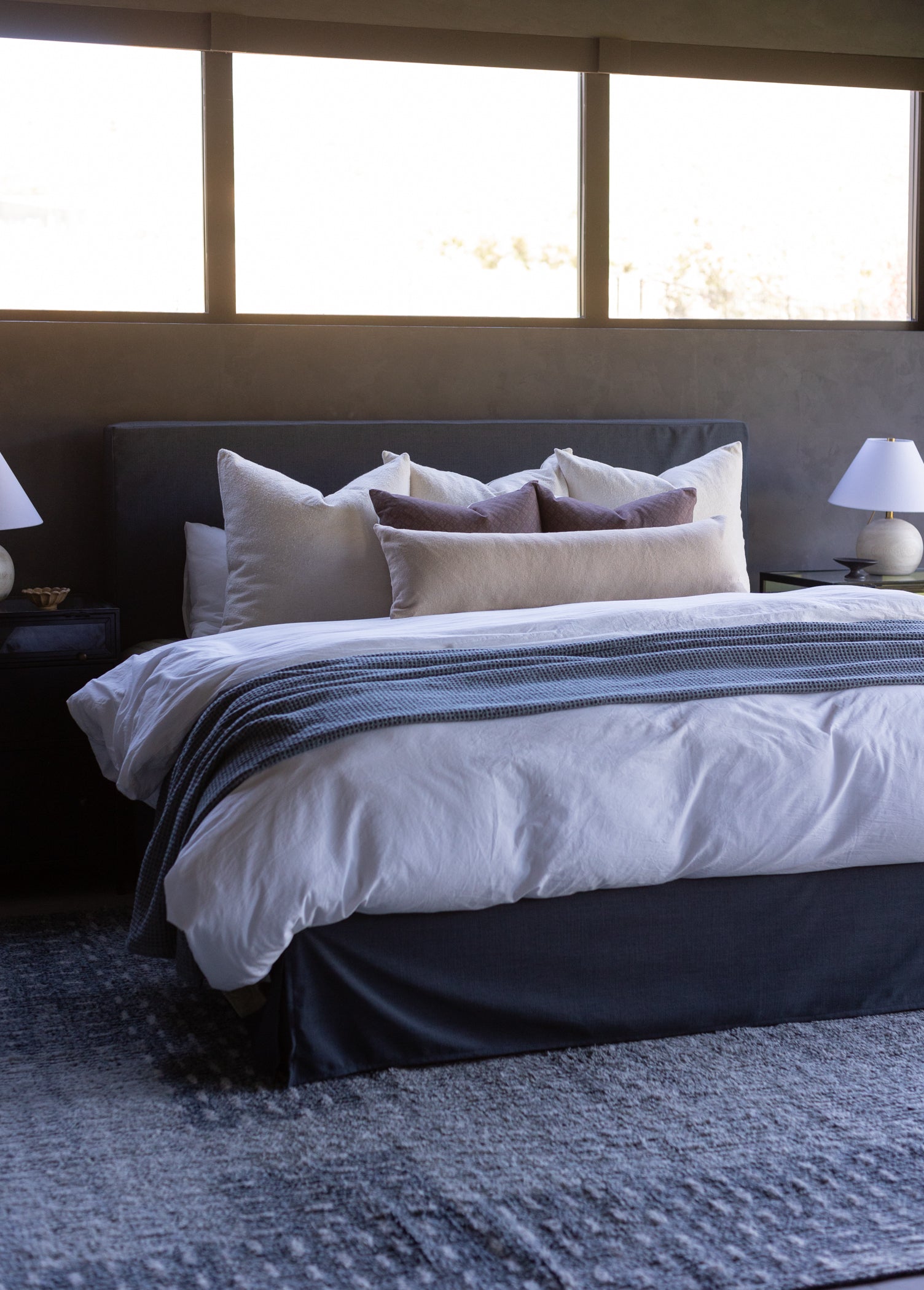 Beige boucle textured pillows arranged on a dark headboard with white bedding and striped throw.