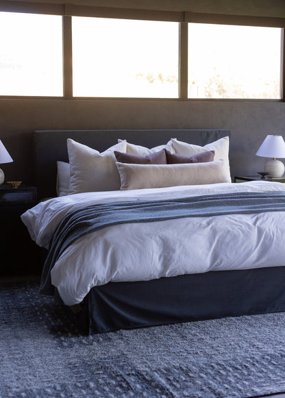 Beige boucle textured pillows arranged on a dark headboard with white bedding and striped throw.