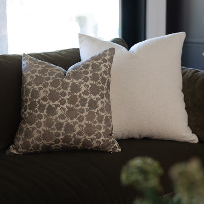 Two throw pillows on dark sofa: one with taupe damask pattern, one cream solid, both square-shaped.