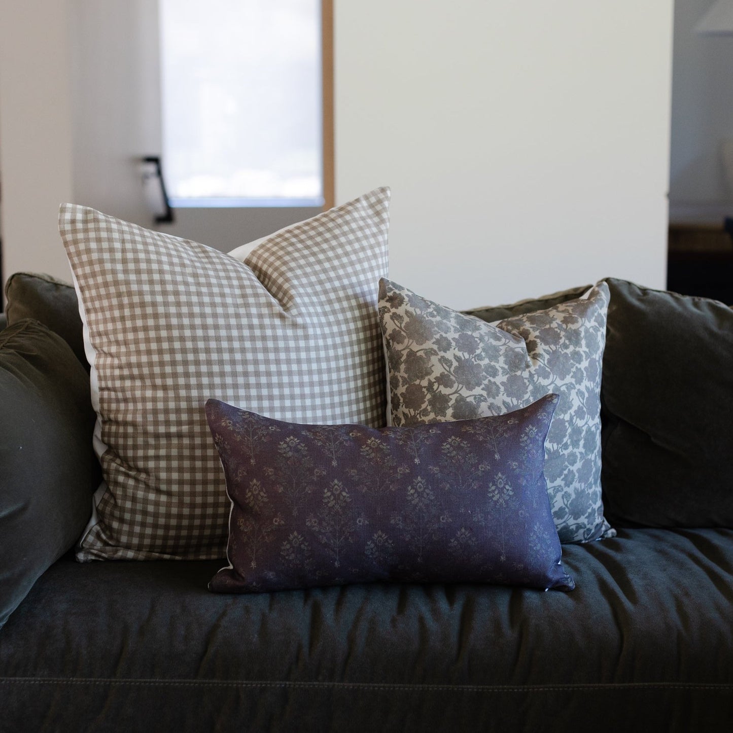 Three decorative pillows on dark sofa: gingham, floral, and damask patterns in neutral and navy tones.
