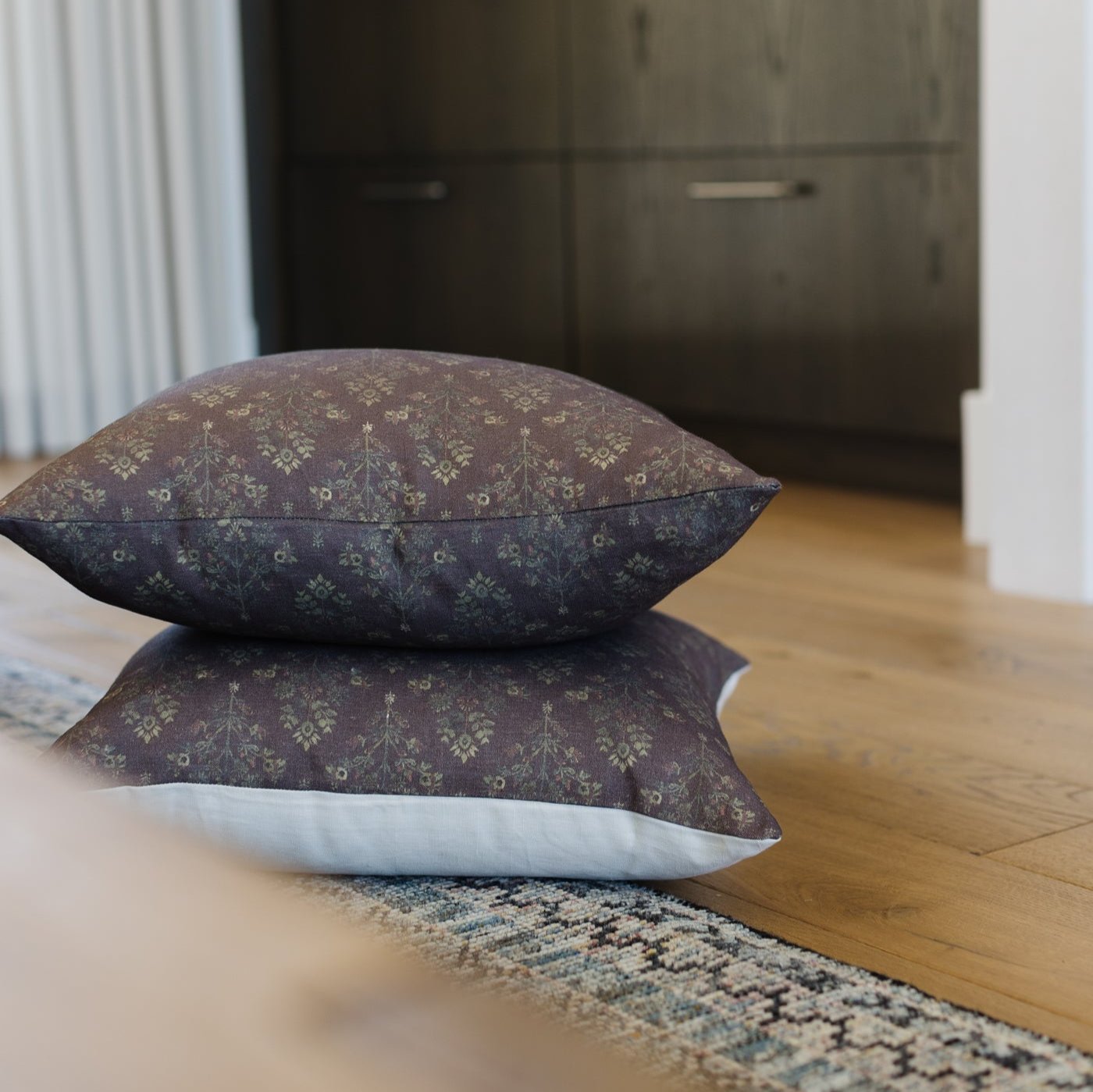 Stacked linen pillows in burgundy and navy with geometric damask pattern on wooden floor.