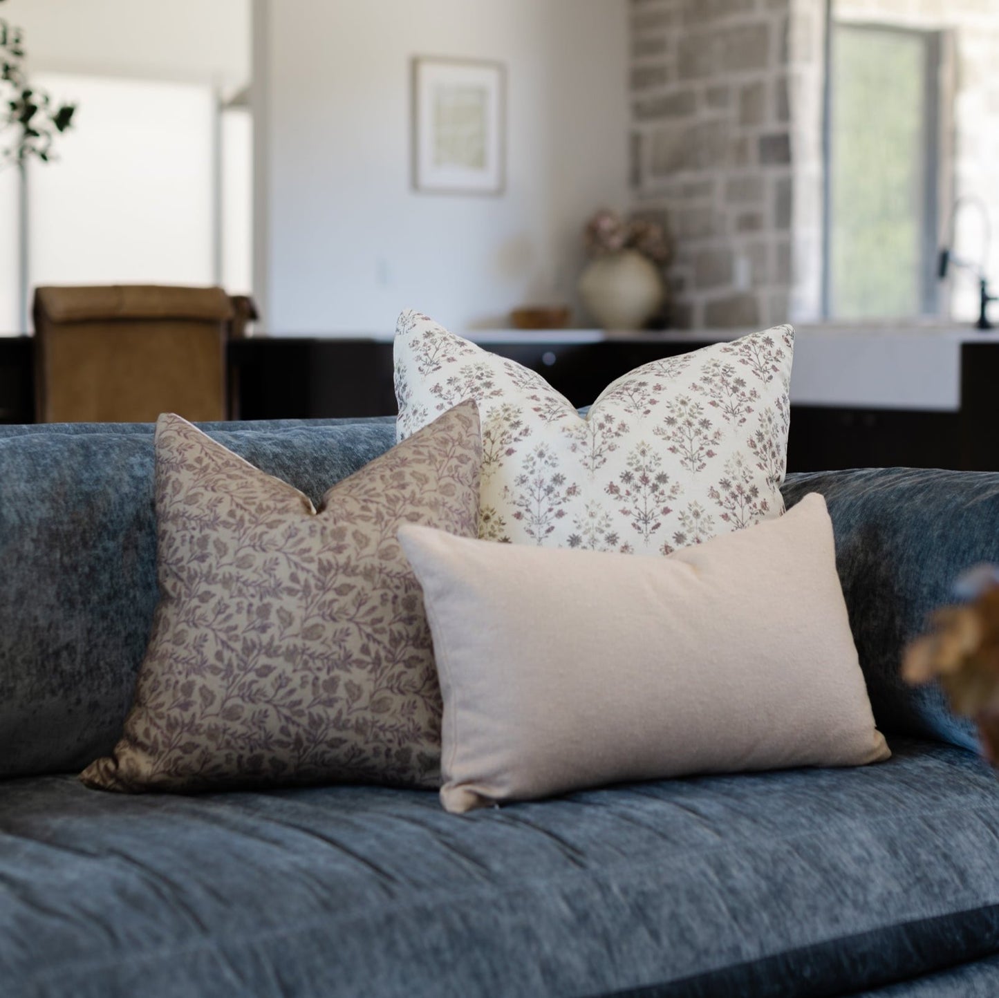 Three decorative pillows in neutral tones displayed on a blue-gray sofa in a bright living room.