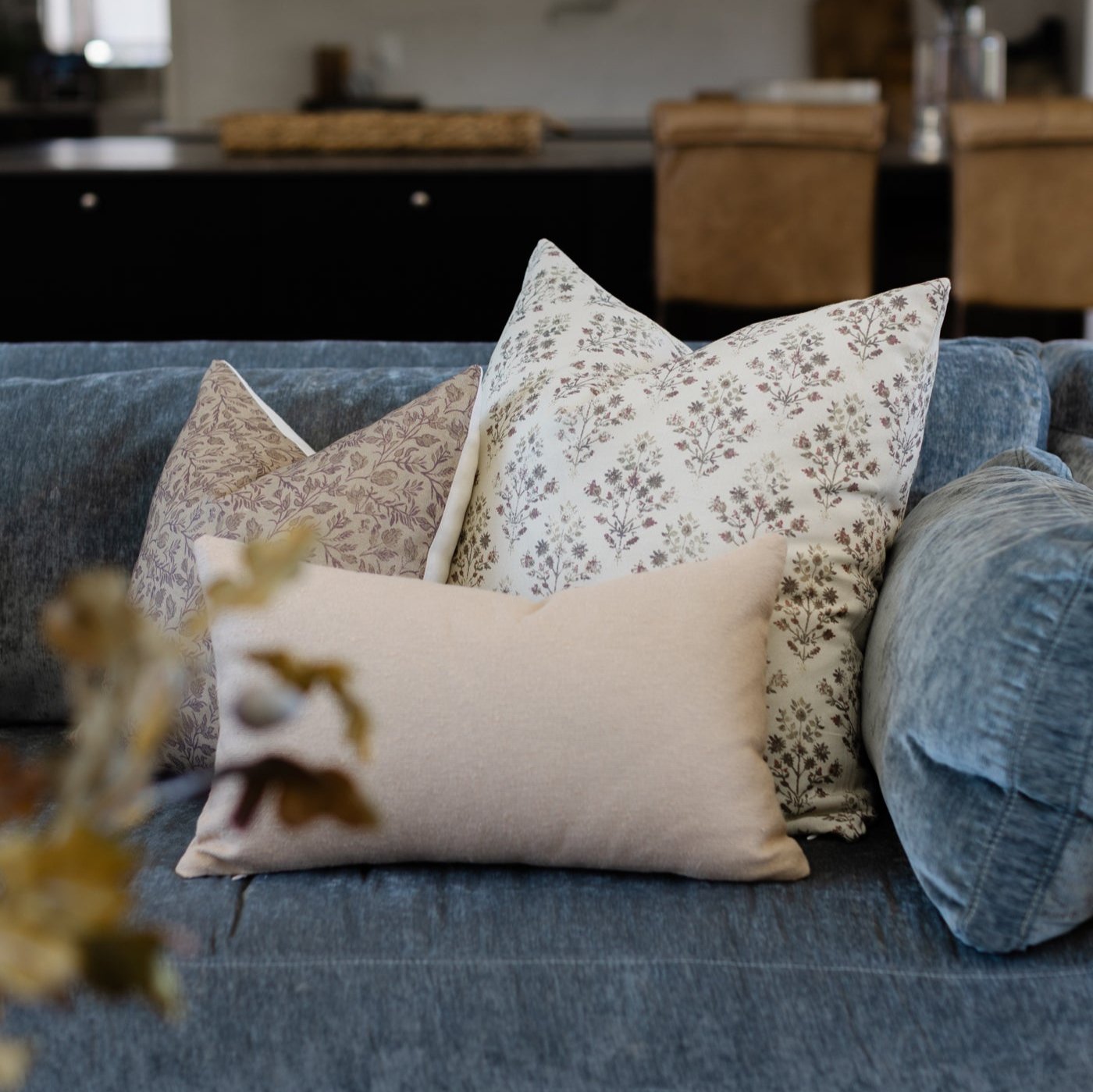 Three decorative pillows on a blue upholstered sofa: cream solid, taupe floral, and cream floral with leaf pattern.