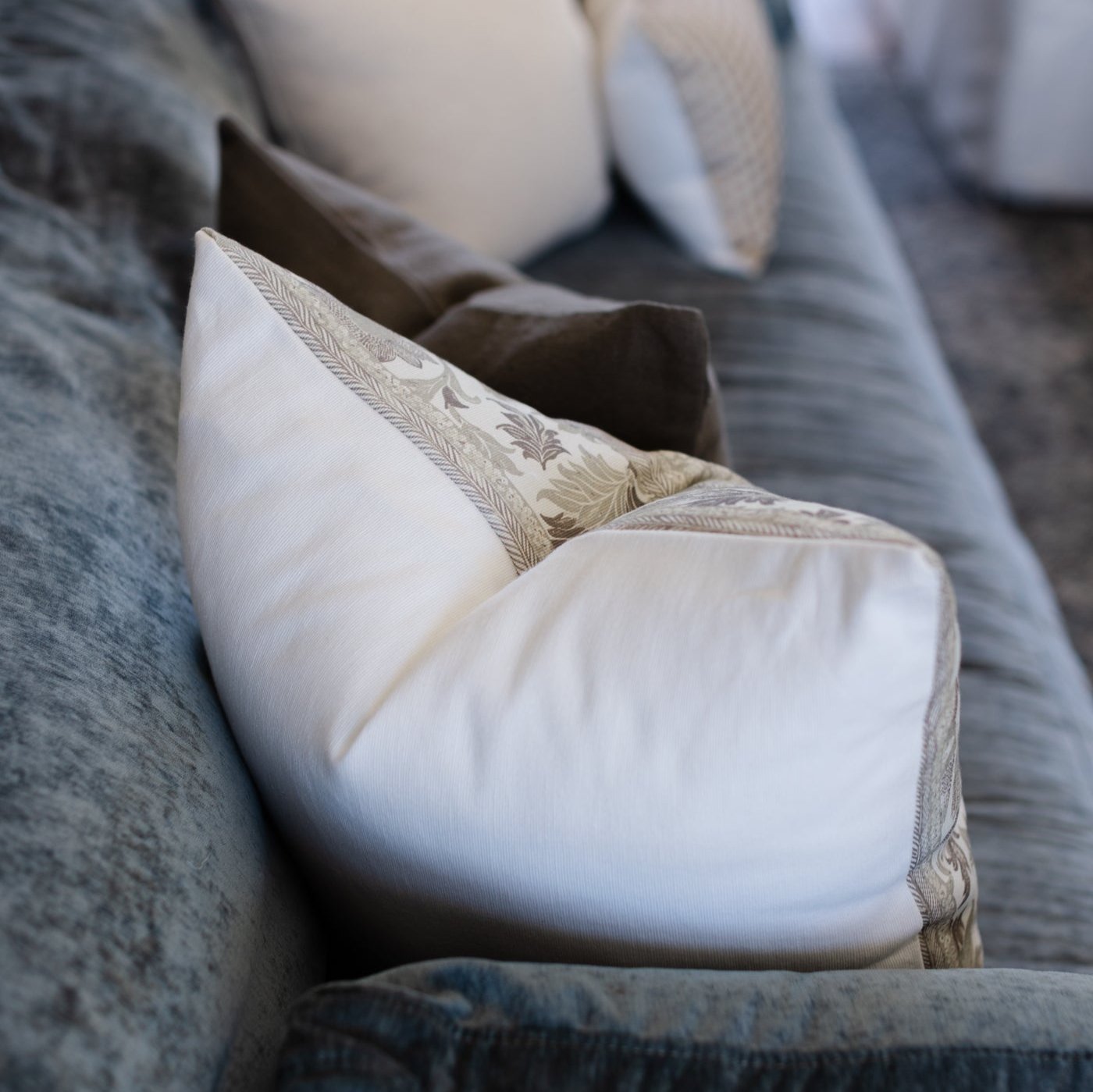White and cream decorative pillows with patterned trim arranged on gray corduroy upholstered furniture.