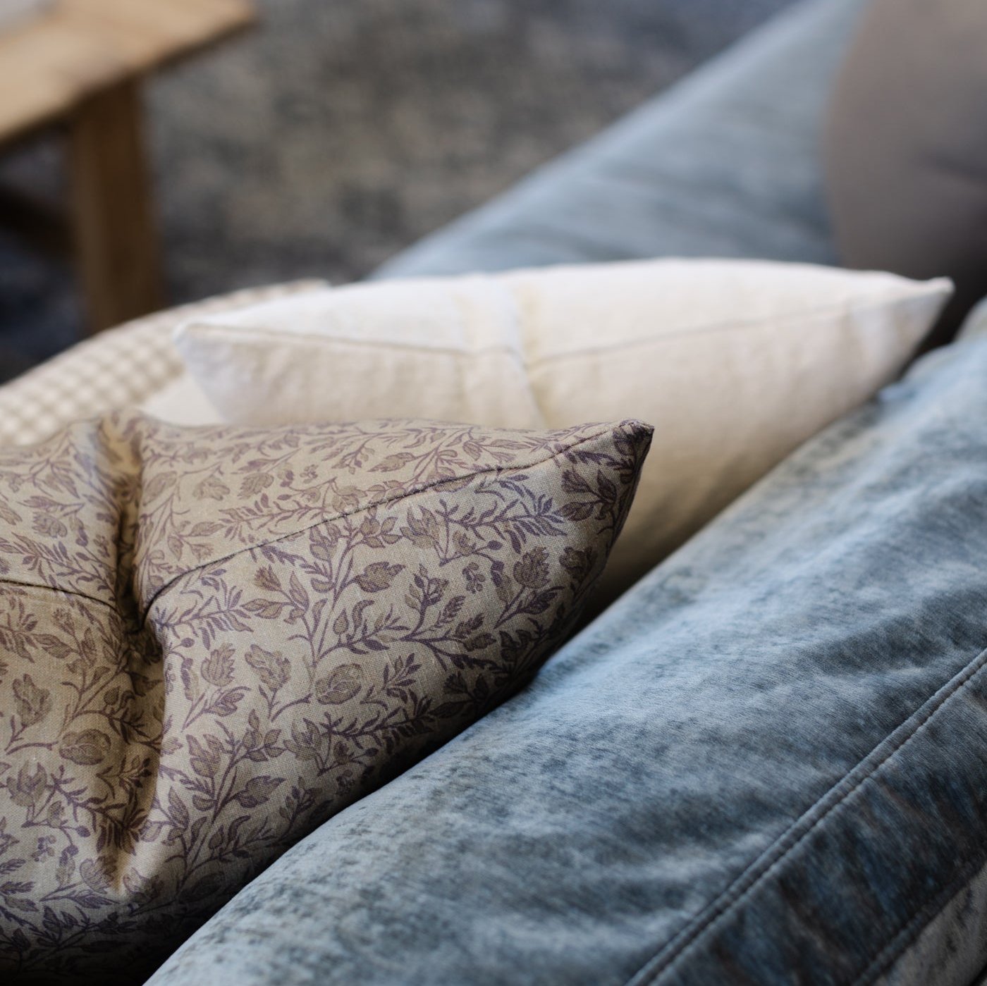 Decorative pillows in taupe floral, cream textured, and cream check patterns arranged on blue upholstered furniture.