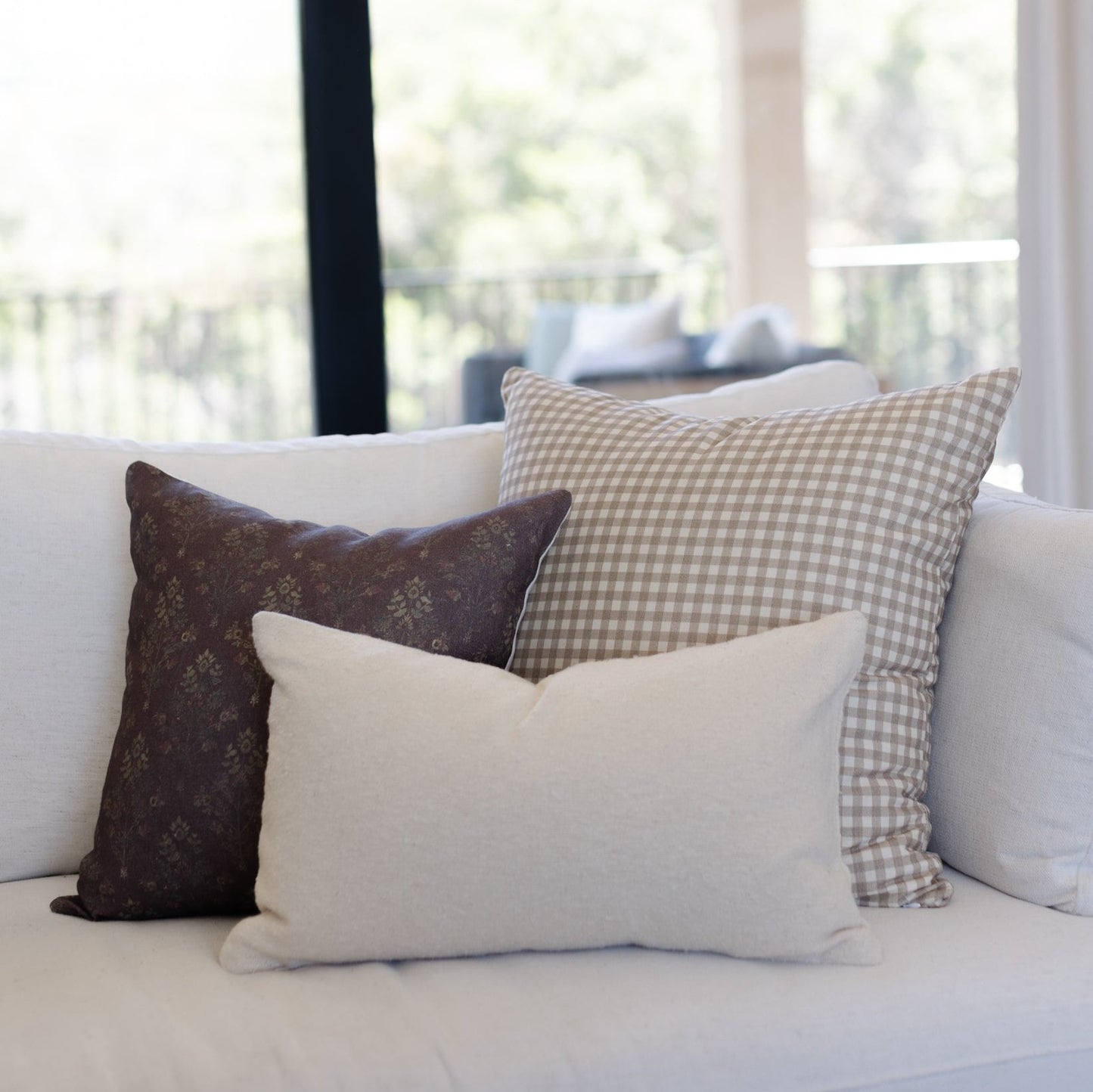 Three decorative pillows arranged on a light sofa: dark burgundy floral, cream solid, and taupe gingham patterns.