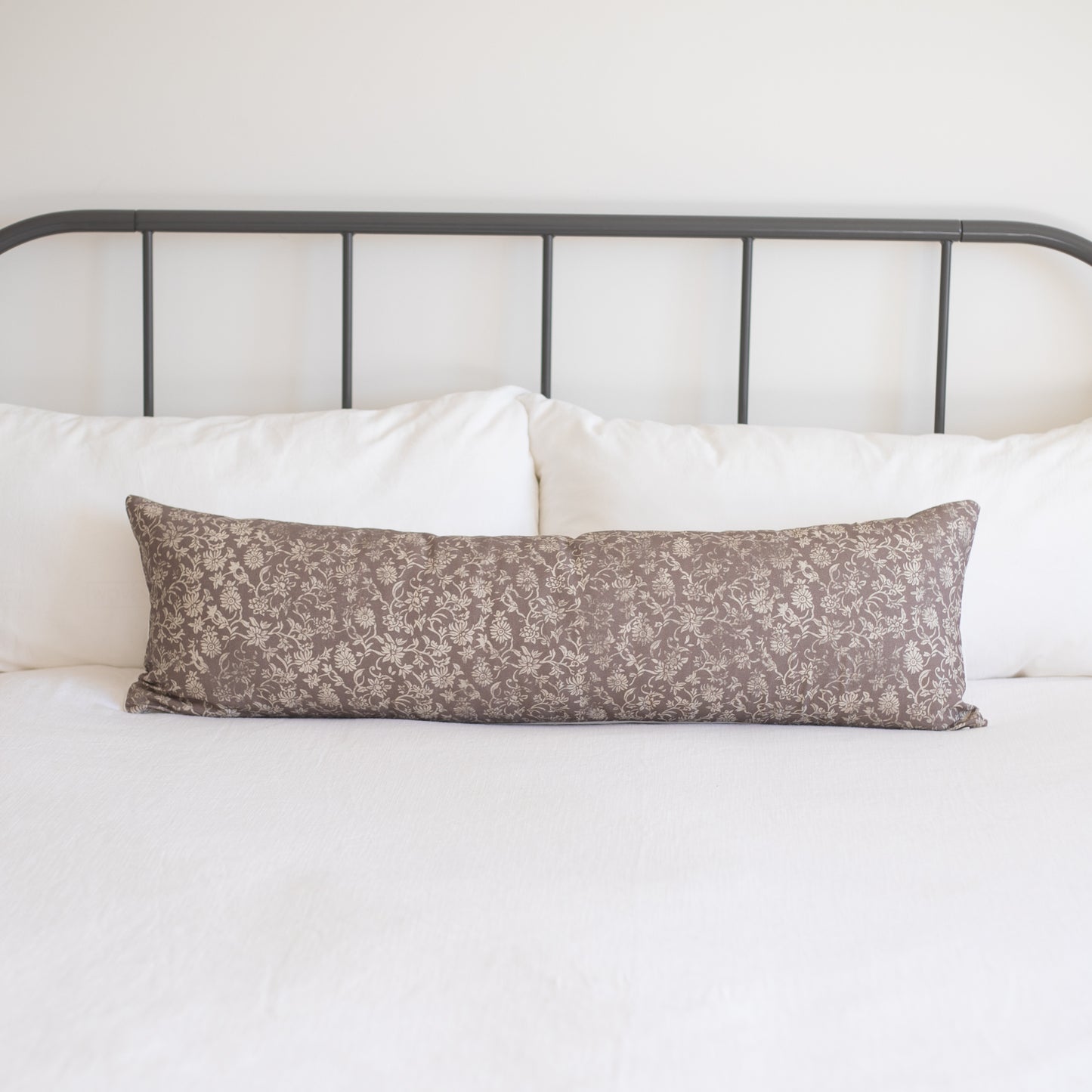 Taupe lumbar pillow with cream floral damask pattern displayed on white bedding against metal headboard.
